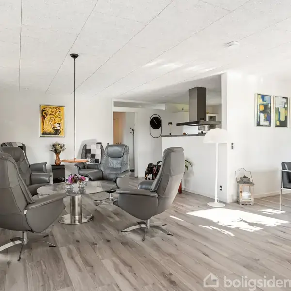 Grå lænestole er placeret rundt om et glasbord med blomster, i en lys stue med trægulve. Der er kunst på væggene og en spiseplads i baggrunden.