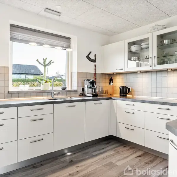 Hvidt køkken med L-formet bordplade, kaffemaskine og elkedel på bordet. Store vinduer giver udsigt til have og træ. Lysegrå fliser langs væggen. Laminatgulv.