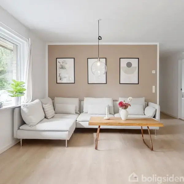 En lysegrå sofa står mod en beige væg med tre indrammede billeder, akkompagneret af et træsofabor med en vase med blomster. Et stort vindue til venstre lader lys strømme ind.