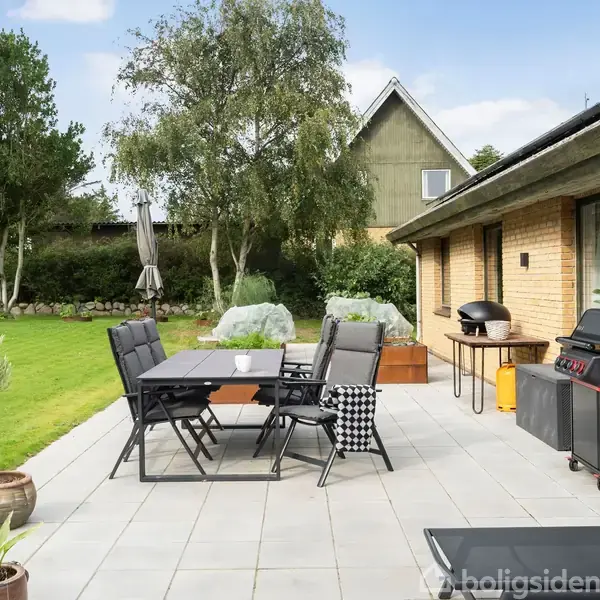 Terrassemøbler står på en flisebelagt terrasse ved et gult murstenshus. Grill og spisebord er placeret blandt krukker og planter i en grøn have med træer i baggrunden.
