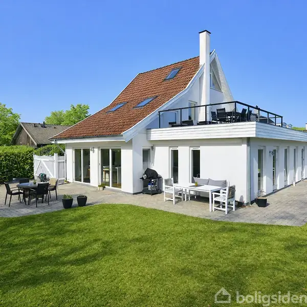 En moderne, hvid villa med rødt tegltag står i en grøn have. Flere terrassemøbler er placeret på fliserne rundt om huset. Skyfri blå himmel markerer solrig dag.