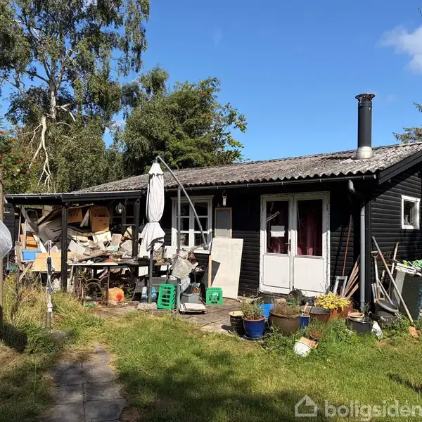 Et sort træhus står i en have med frodig vegetation. Udendørs er der mange ting som paraply, krukker, og byggematerialer, hvilket skaber et rodet indtryk.