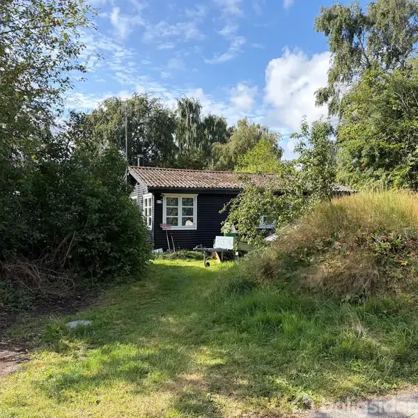Et sort træhus står stille omgivet af frodige træer og grønne buske. Foran huset ses en græsplæne og en mindre bakke, under en blå skyet himmel.