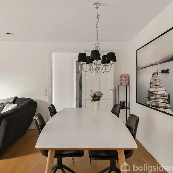 Spisebord omgivet af sorte stole står i en stue med hvidmalede vægge. En sort-hvid lysekrone hænger over bordet. Til højre ses et stort billede af en mole med båd i baggrunden.