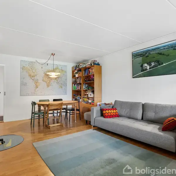 En grå sofa står i en stue med træmøbler. Et kort hænger på væggen over et spisebord med fire stole. En bogreol og lamper skaber en hyggelig atmosfære.
