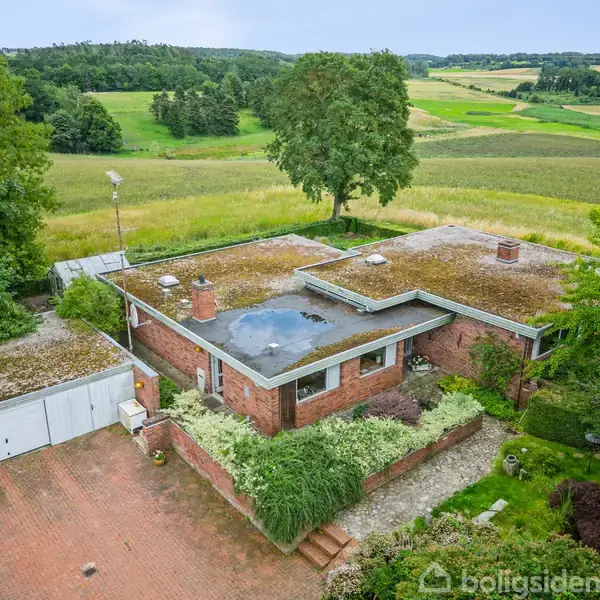 En lav murstensvilla med fladt tag ligger omgivet af velholdt have. En stor gårdsplads er foran huset, og landskabet omkring er en frodig landlig udsigt med marker og træer.
