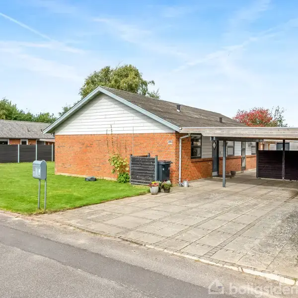 En murstensbungalow med en carport står på en pæn grund. Huset har en velplejet græsplæne og flisebelagt indkørsel. Det ligger i et roligt boligområde med andre huse omkring.