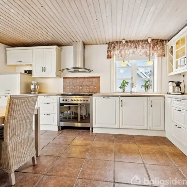 Et lyst køkken med hvide skabe og skuffer. Der er en ovn og kogeplade i midten. Vindue med blomster i baggrunden, spisebord med flettede stole til venstre.