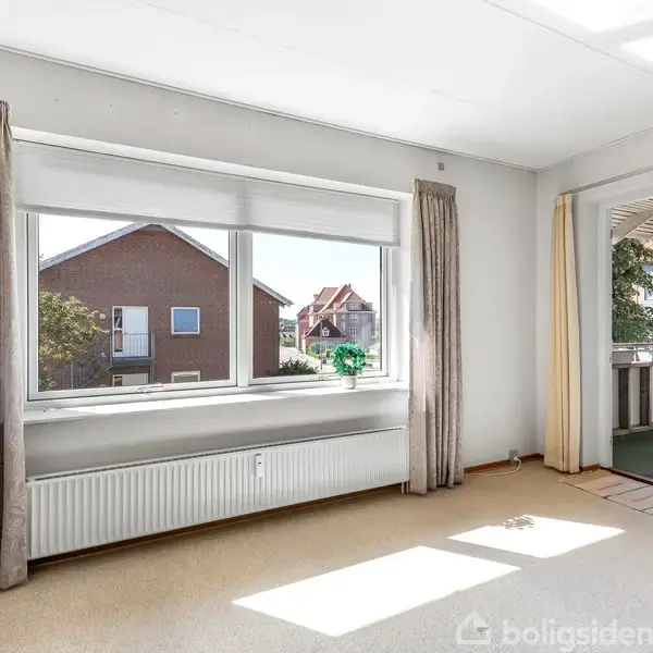 Et lyst værelse med store vinduer og beige gardiner åbner ud mod en balkon. Indenfor er der en radiator under vinduet. Udsigt til nabohuse og blå himmel.