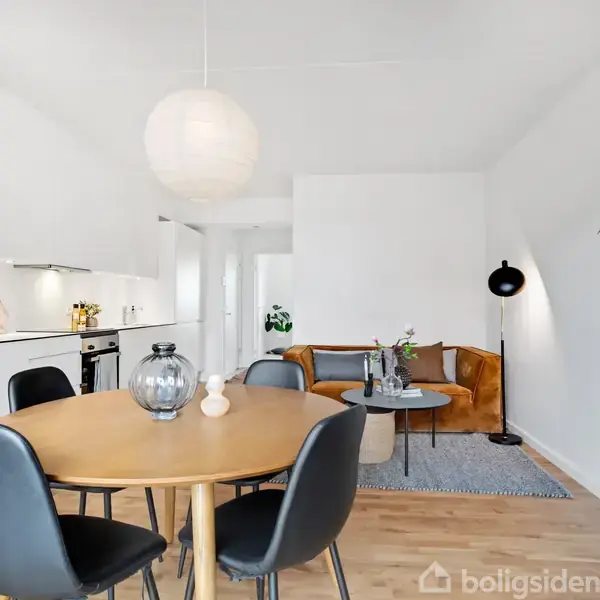 Rundt spisebord med sorte stole står i stuen. I baggrunden ses et lille køkken og en sofa med et sofabord. Et stort indendørs plante skaber en hyggelig atmosfære.