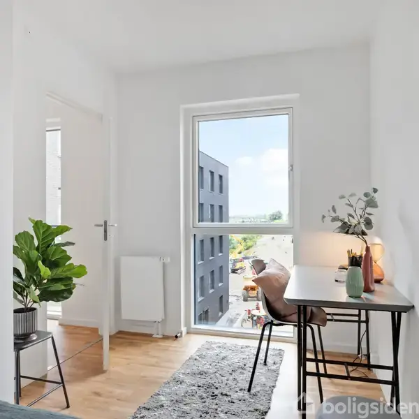 Et skrivebord med en stol og planter står ved et vindue. Rummet er lyst med hvidmalede vægge, trælaminatgulv og udsigt til en bygning udenfor. En dør står åben.