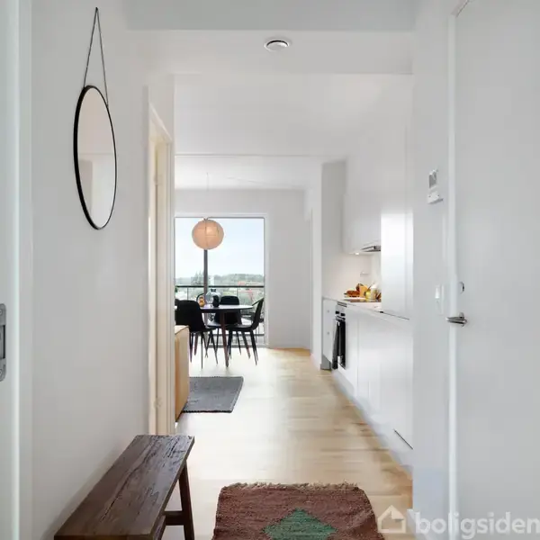 Spejl hænger på en hvid væg i en gang, hvor en bænk står modsat. Gangen fører til et køkken-alrum med spisebord, store vinduer og udsigt over landskabet.