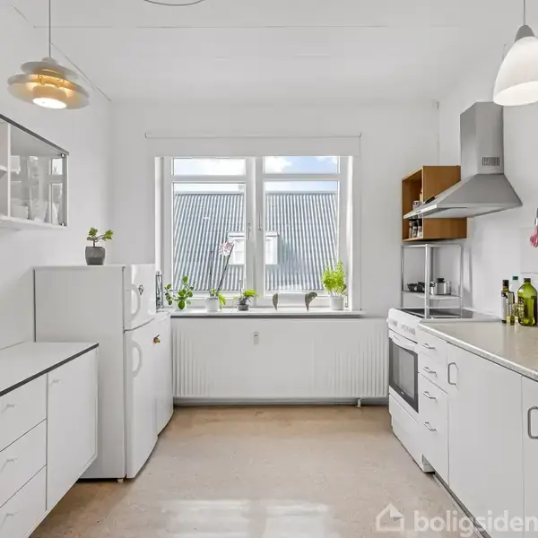 Et hvidt køkken med åbne hylder, køleskab, komfur og knivholder. Vindue med udsigt til taget, omgivet af grønne planter. Lyse lamper hænger fra loftet.