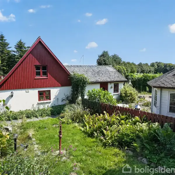 Rødt, bindingsværkshus med græs og buske i forhaven, beliggende i et frodigt, grønt landskab under en blå himmel. En mindre, rund bygning er placeret til højre.