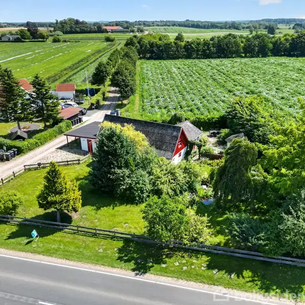 En rød bondegård omgivet af tæt vegetation og træer, set fra luften. Den ligger ved siden af en vej med åbne marker i baggrunden.