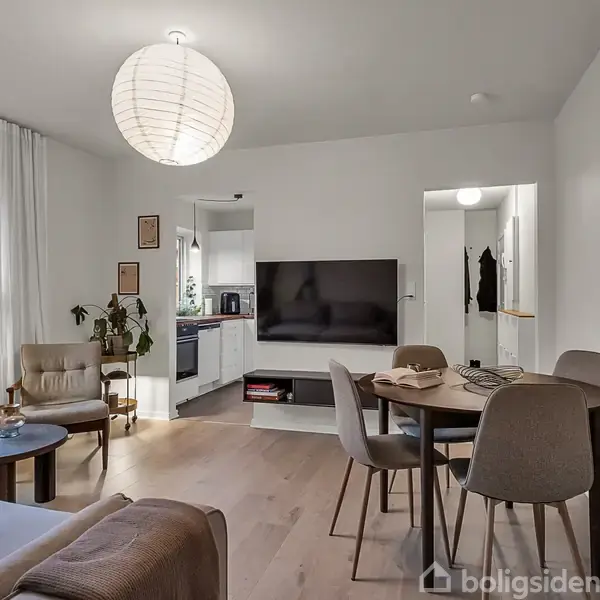 Stue med moderne møbler: En sofa, rund spisebord med stole, og en stor fladskærms-TV på væggen. Køkken i baggrunden, lyst trægulv og vindue mod lysende bygninger udenfor.