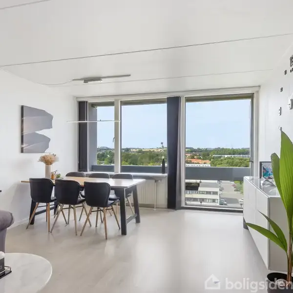 En moderne stue med spisebord og stole ved et stort vindue, der afslører en udsigt over byen. En sofa til venstre, planter og vægdekorationer skaber en hyggelig atmosfære.