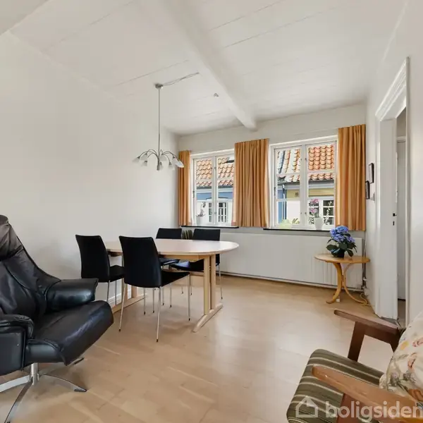 En stue med en sort læderlænestol og et rundt spisebord med fire stole. Baggrunden har store vinduer med gardiner, og en lille sidebord med en blå plante.