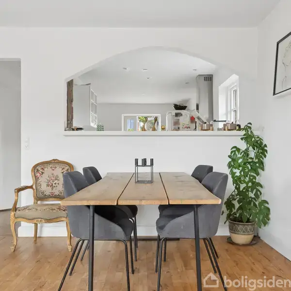 Et spisebord med seks grå stole står på et trægulv, omgivet af hvide vægge; en grøn plante er placeret til højre, og en antik stol står ved siden af en hvid døråbning.
