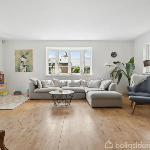 Stue med stor grå hjørnesofa, sofabord og lænestol. Der er lyse trægulve, store vinduer, planter og legetøjshjørne. Kunstmaleri hænger på væggen. Vinduerne udsigt til bygninger.