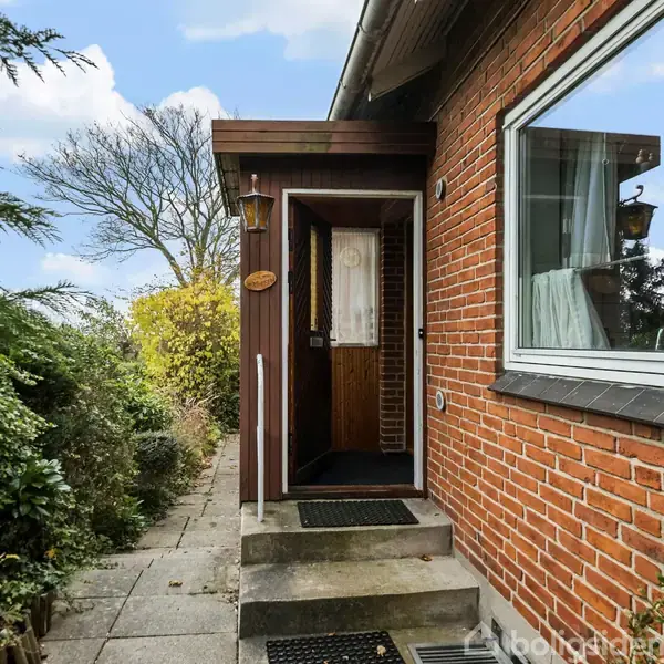 En rød murstensbygning har en indgangsdør med et lille trin foran, omgivet af grønne buske og en flisebelagt sti. Ved siden af døren er et vindue.