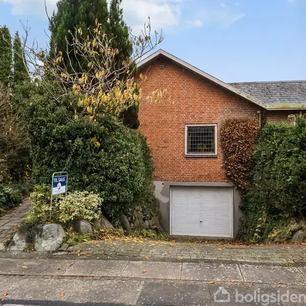Et rødt murstenshus med en garageport og en lille firkantet vindue, omgivet af frodig vegetation. Til salg-skilt tæt ved indgangstrappen.