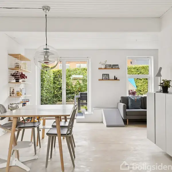 En lys spisestue med et spisebord og stole i træ, beliggende foran et stort vindue med udsigt til en grøn have. Der er hylder på væggen og en sofa i baggrunden.