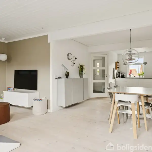 Stue med gråt sofaarrangement og brun puf ved siden af et minimalistisk TV-møbel på en beige væg. Rundt spisebord med stole nær hvidt køkken under glaslampe. Indbydende, moderne atmosfære.