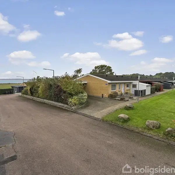 Et gulstenshus står ved siden af en grusvej omgivet af grønne græsarealer og buske. En bil ses i forgrunden, og marker strækker sig i baggrunden under en blå himmel.