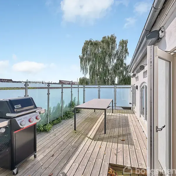Grill står på træterrasse ved siden af et bord. Omgivet af glasrækværk under en blå himmel, med udsigt til vand og træer. Terrasse er ved et hus med åben dør.