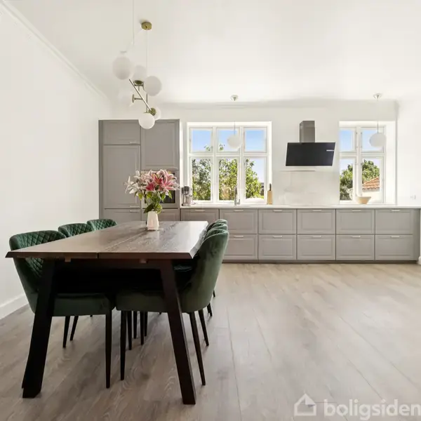 Spisebord med grønne stole står i et lyst, rummeligt køkken-alrum med trægulv. Grå køkkenskabe og hvide vægge omkranser store vinduer. Vase med blomster pryder bordets midte.