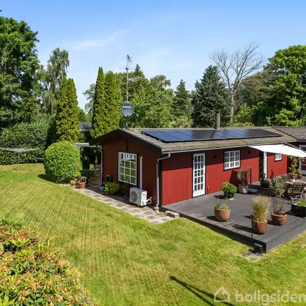 Rødt hus med fladt tag og solpaneler står i en frodig have. Terrasse med krukkeplanter ved huset. Omgivet af grønt græs og høje træer.