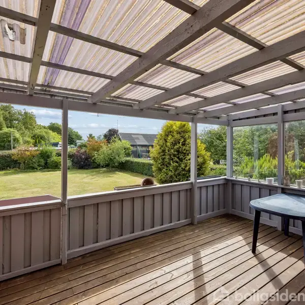 En overdækket terrasse med et gennemsigtigt tag har en tom firkantet bordplade. Udsigt til en stor grøn have med forskellige buske og et hus i baggrunden.