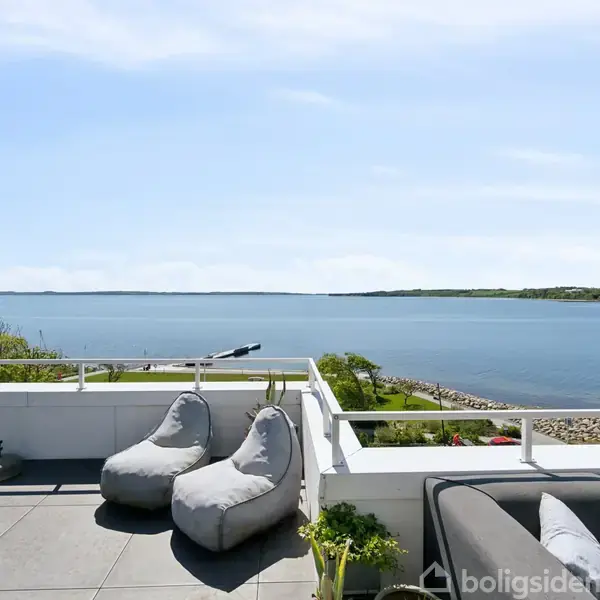 Terrassemøbler, herunder to grå sækkestole og en sofa, står stille på en flisebelagt terrasse med udsigt over en stille bugt og landskab i baggrunden.
