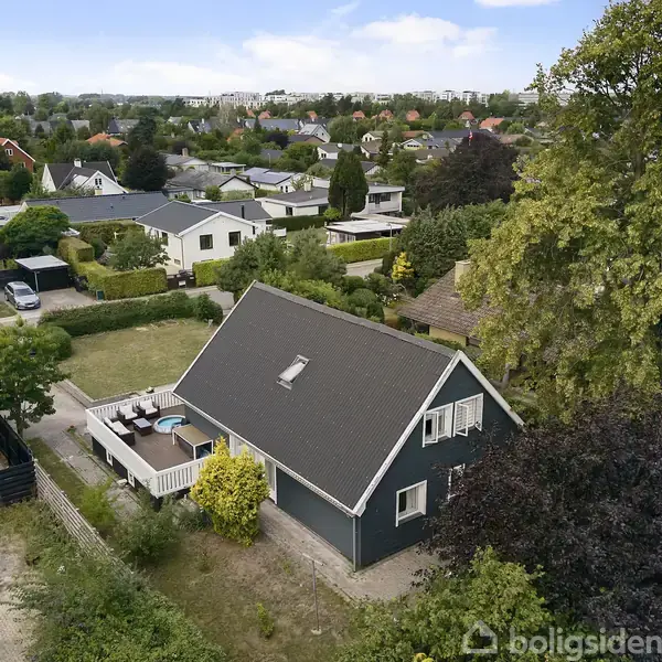 Et sort træhus med skråt tag og kvist står i en have. Haven har terrasse med møbler. Huset er omgivet af tæt bebyggede parcelhuse og træer i et villakvarter.