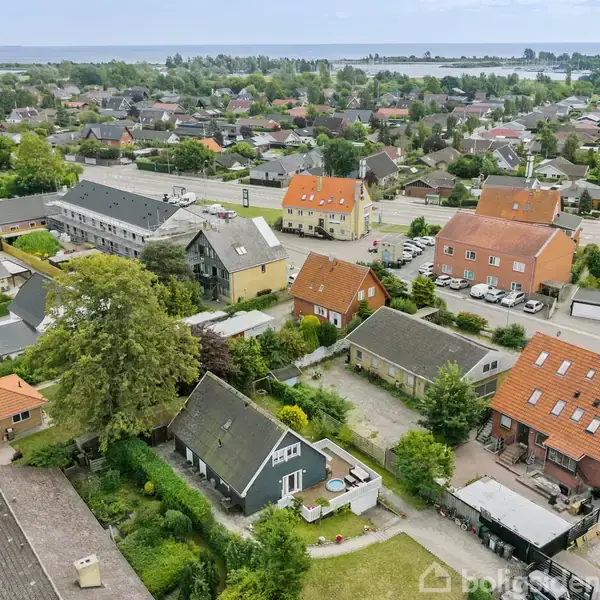 Luftfoto af en by med rækkehuse med orange tage, træer og haver omkring. Flere større bygninger ses i baggrunden, og en sø er synlig i horisonten.