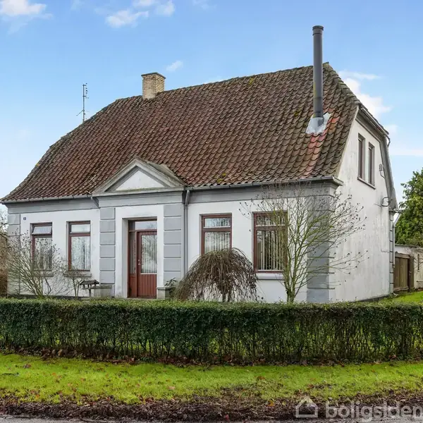 Et hvidmalet hus med rødt tegltag står på en grøn græsplæne omgivet af hække. To etager med flere vinduer, omgivet af træer og buske i et landligt område.