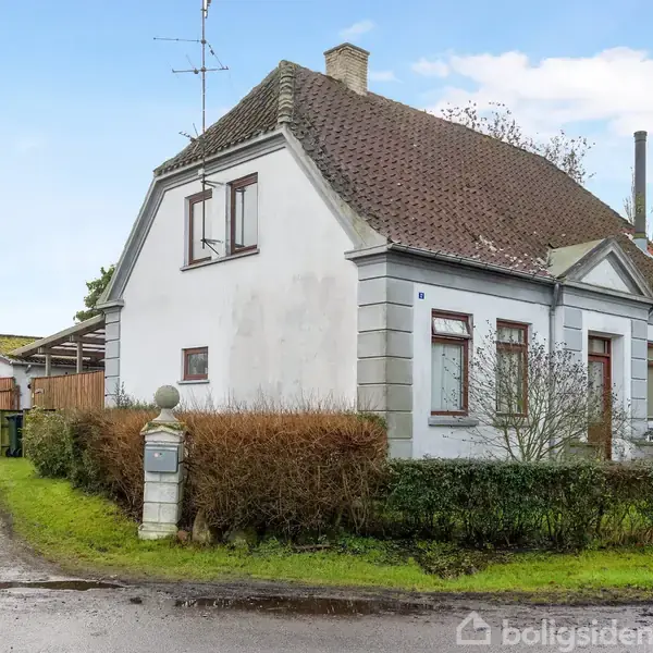 Et hvidt hus med rødt tegltag står stille på en hjørnegrund omgivet af grønne buske og en grusvej. I baggrunden ses en trægarage.