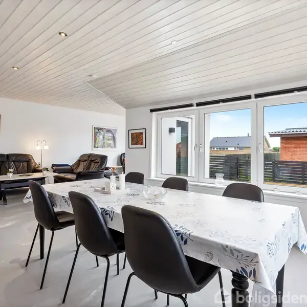 Spisebord med seks stole står i en stue med lyse vægge. Store vinduer giver udsigt til en have og nabobygning. Sofagruppe og dekoreret væg i baggrunden.