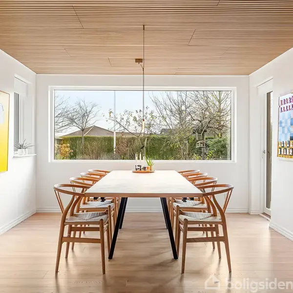 Et stilfuldt spisebord med otte træstole står i et lyst rum. Bordet har en lys dekoration, og store vinduer tilbyder udsigt til en have. Farverige malerier hænger på væggene.