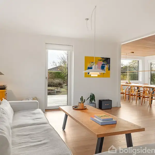 En stue med en hvid sofa og et træbord centreret foran et stort fladskærms-tv. En farverig plakat hænger på væggen. I baggrunden ses et spisebord ved vinduer.