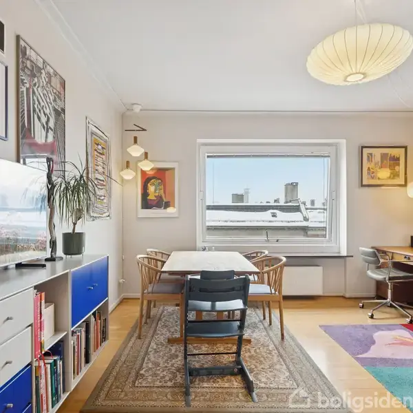 Stue indrettet med et spisebord og stole midt i rummet, omgivet af farverig kunst på væggene. En stor lampe hænger fra loftet, ved siden af et skrivebord.