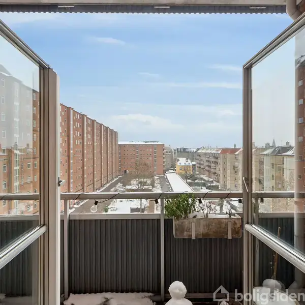 En altan med åbne glasdøre vender ud mod en udsigt over en bymæssig scene med røde murstensbygninger. Sne dækker jorden, og en plante hænger på rækværket.