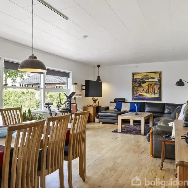 Spisestue med træbord og stole, opholdsstue med sort sofa og vægkunst, store vinduer giver udsigt til have.