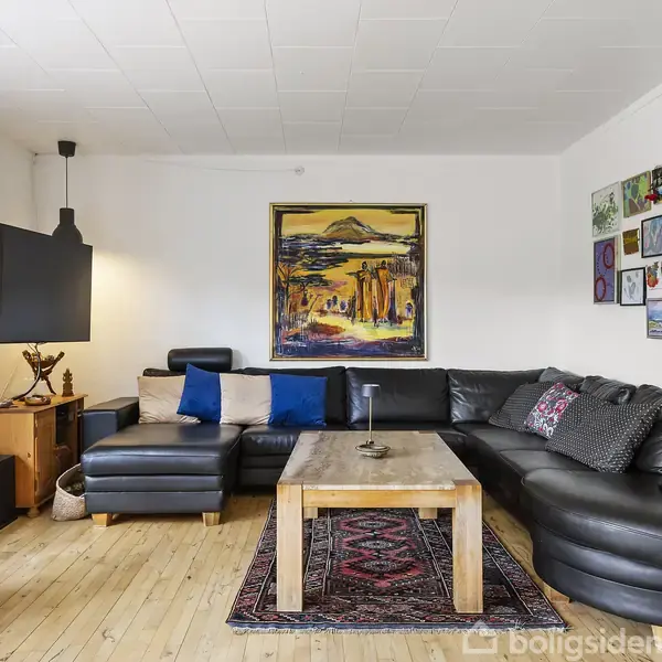 Sort lædersofa står i hjørnet af en stue med trægulv, flankeret af kunst på væggene. En fladskærm hænger ved siden af en standerlampe. En træstuebord står centralt.