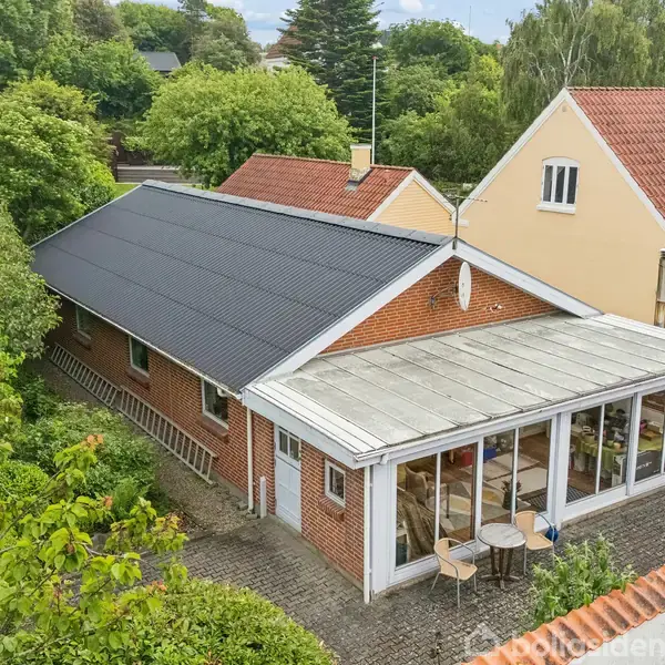 En rød murstensbygning med hvide vinduer og en hvid hoveddør står langs en brolagt sti. Der er planter i krukker langs facaden, og en træhegn begrænser stien.