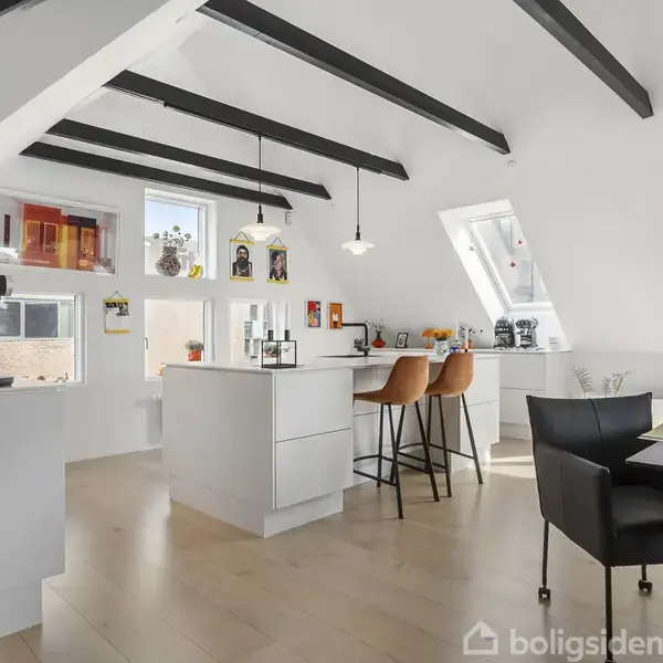 Køkken i loftslejlighed har hvide skabe og barstole ved køkkenø. Skråvinduer og spær i loftet skaber lys og rummelighed. Spiseplads med bord og stole ved siden af.