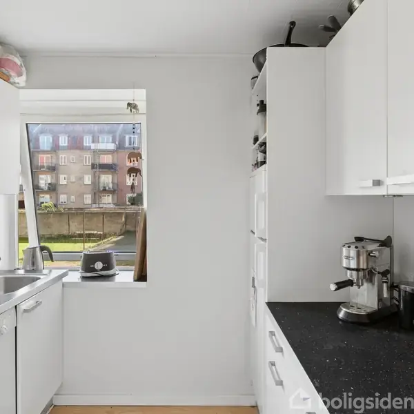 Et smalt køkken med hvide skabe og sort bordplade. En kaffemaskine og knivblok står på bordet. Et vindue giver udsigt til en boligbygning udenfor.