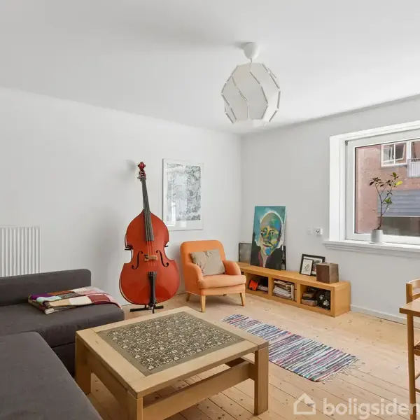 En stue med en kontrabas ved siden af en orange stol. Et gråt hjørnesofaarrangement står tæt ved et lavt sofabord. Der er træmøbler, en reol og et stort vindue.