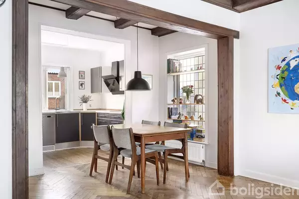 Et spisebord med fire stole står i et lyst rum ved siden af en åben køkkenindgang med moderne skab og dekorationer. En bogreol og farverigt kunstværk pryder væggene.